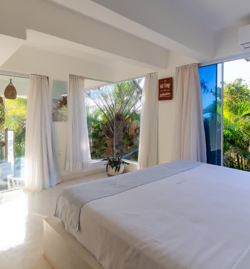 Kilombo Villas - villa Zumbi Jardim - Quarto amplo e iluminado da villa Zumbi Jardim com paredes brancas e cama king size. Grandes janelas de vidro do chão ao teto oferecem uma vista exuberante do jardim tropical, com cortinas brancas fluidas e uma placa decorativa onde se lê "No time like paradise".