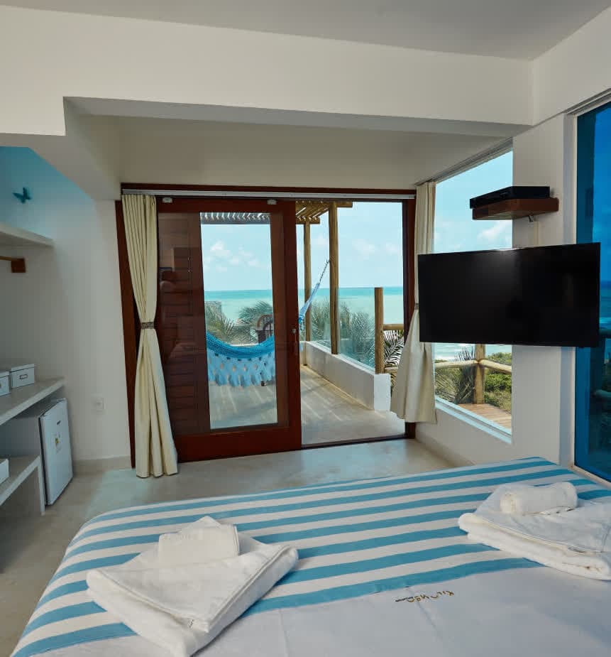Quarto iluminado com cama de casal decorada com colcha listrada em azul e branco. Ao fundo, uma porta de correr de madeira abre para uma varanda com rede azul e vista panorâmica para o mar. Uma televisão de tela plana está fixada na parede lateral.