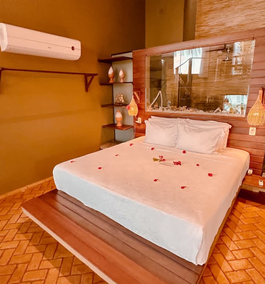 Kilombo Villas - villa Desidéria - Imagem de um quarto de hotel com decoração moderna, cama com lençol branco e flores, painel de madeira atrás da cama e prateleiras decorativas ao lado.