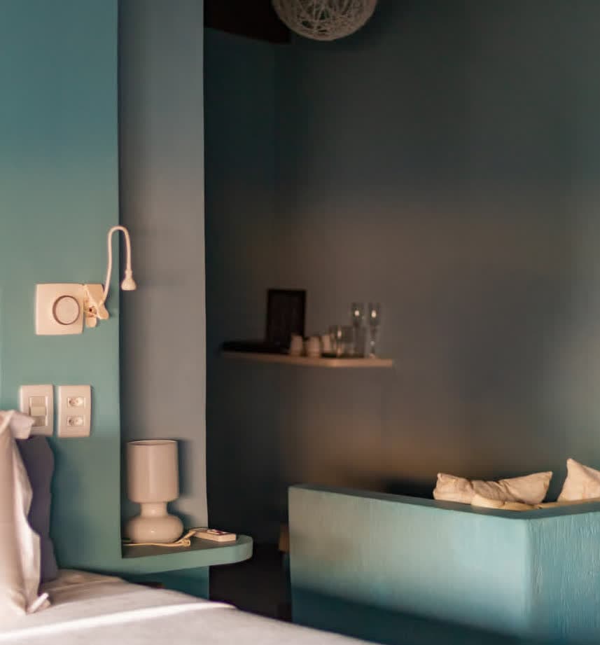 Kilombo Villas - Suíte Azul - Detalhe da cabeceira na Villa Suíte Azul, com paredes em tom azul-celeste. O espaço inclui uma luminária de leitura articulada, interruptores modernos e uma mesa de cabeceira integrada, além de uma vista parcial de uma área de estar rebaixada ao fundo.