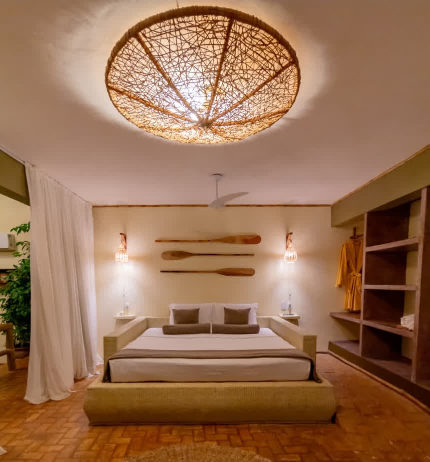 Kilombo Villas - villa Sibraáma Jardim - Quarto principal da villa Sibraáma Jardim com foco em uma cama de casal sobre uma base de fibra natural. A decoração é rústica, apresentando remos de madeira decorativos na parede, luminárias pendentes de palha e um grande lustre circular de tramas naturais no teto.