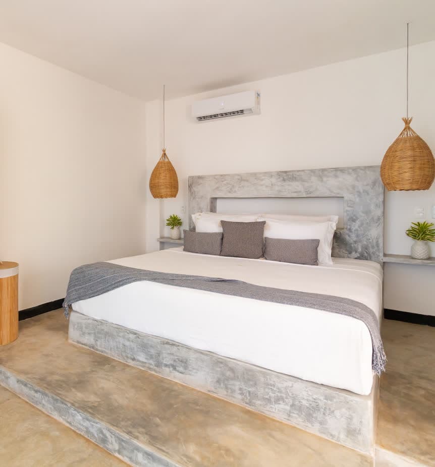 Quarto master com design minimalista em cimento queimado. Uma cama king size está posicionada sobre uma plataforma elevada de concreto, com cabeceira integrada no mesmo material. Luminárias de palha trançada e um cesto de bambu complementam o estilo rústico-contemporâneo.