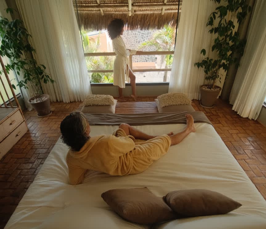 Kilombo Villas - Sibraáma Ocean - Vista de cima do quarto integrado à varanda. Um homem de roupão amarelo está deitado na cama, enquanto uma mulher de roupão branco observa a vista da varanda. O piso é de tijolos aparentes e o teto da varanda é de palha.