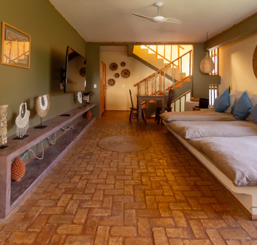Kilombo Villas - Sibraáma Ocean - Ampla sala de estar da villa com piso de tijolos rústicos dispostos em espinha de peixe. À esquerda, uma bancada comprida exibe objetos decorativos étnicos. Ao fundo, vê-se a mesa de jantar e a escada de bambu que leva ao andar superior.