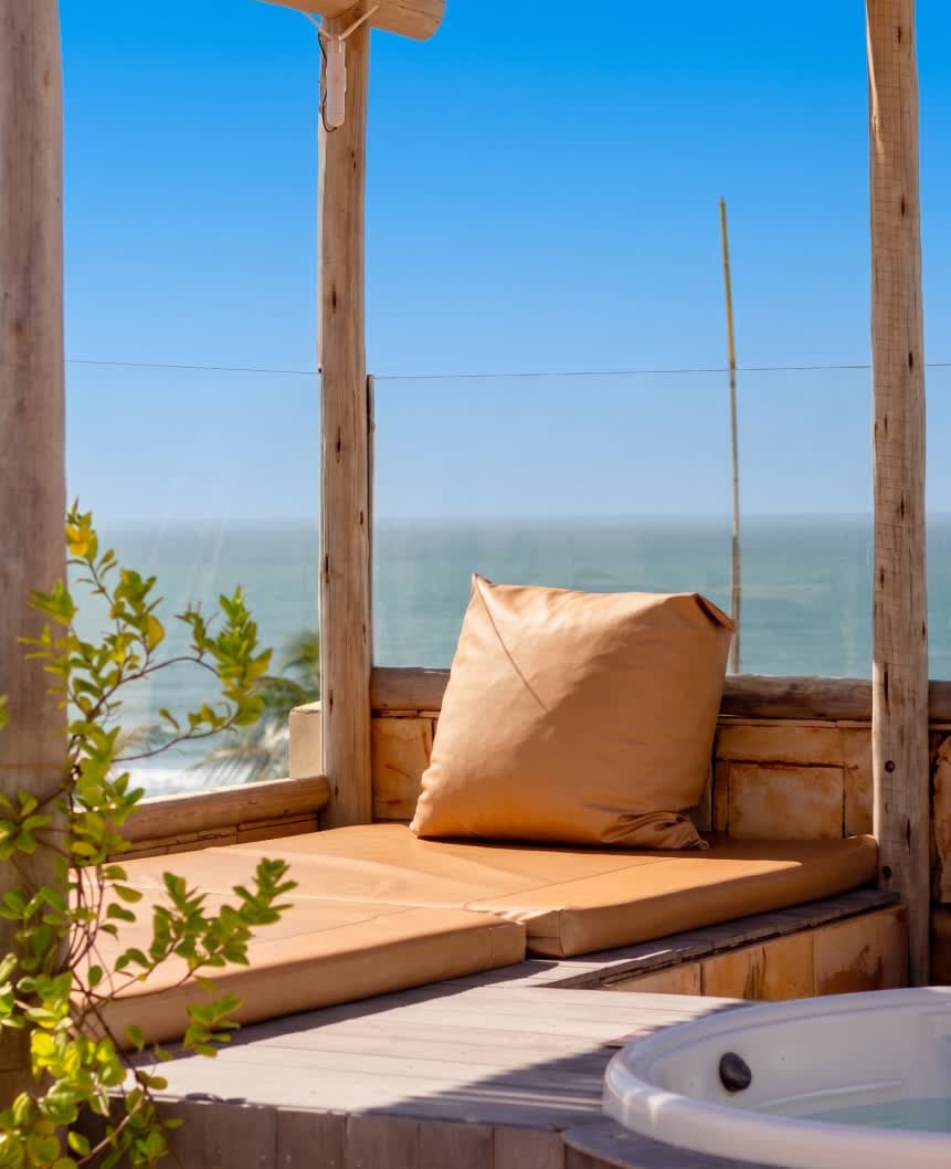 Kilombo Villas - Sibraáma Ocean - Detalhe de uma espreguiçadeira estofada em couro caramelo localizada em um solário privativo. Ao fundo, a linha do horizonte encontra o mar azul sob um céu limpo, emoldurada por pilares de madeira rústica.