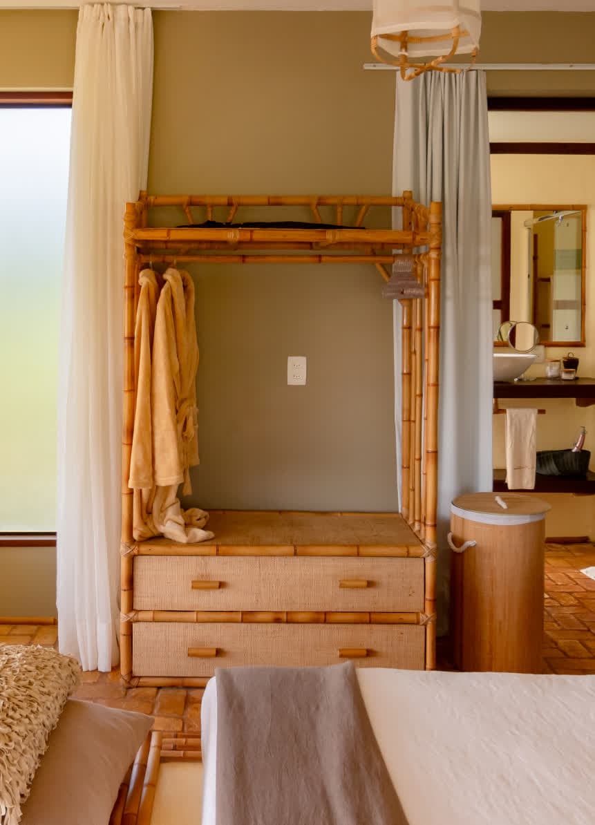Kilombo Villas - Sibraáma Ocean - Mobiliário funcional feito inteiramente de bambu, funcionando como arara de roupas e gaveteiro com acabamento em palha trançada. Roupões felpudos estão pendurados, e a cortina branca ao lado suaviza a entrada de luz.