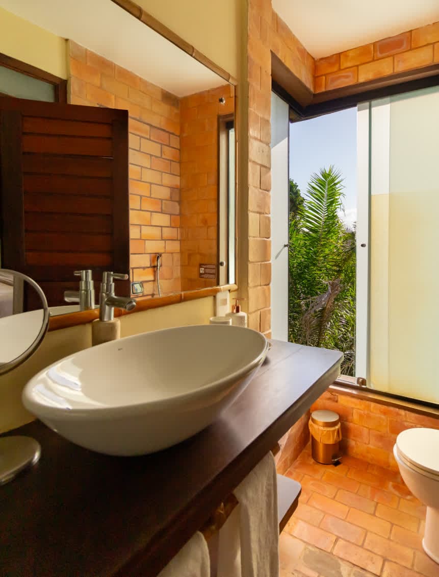 Kilombo Villas - Sibraáma Ocean - Banheiro com revestimento de tijolos aparentes e uma grande janela que oferece vista para a vegetação tropical. A bancada de madeira escura e a cuba branca mantêm o estilo rústico e sofisticado da villa Sibraáma Ocean.