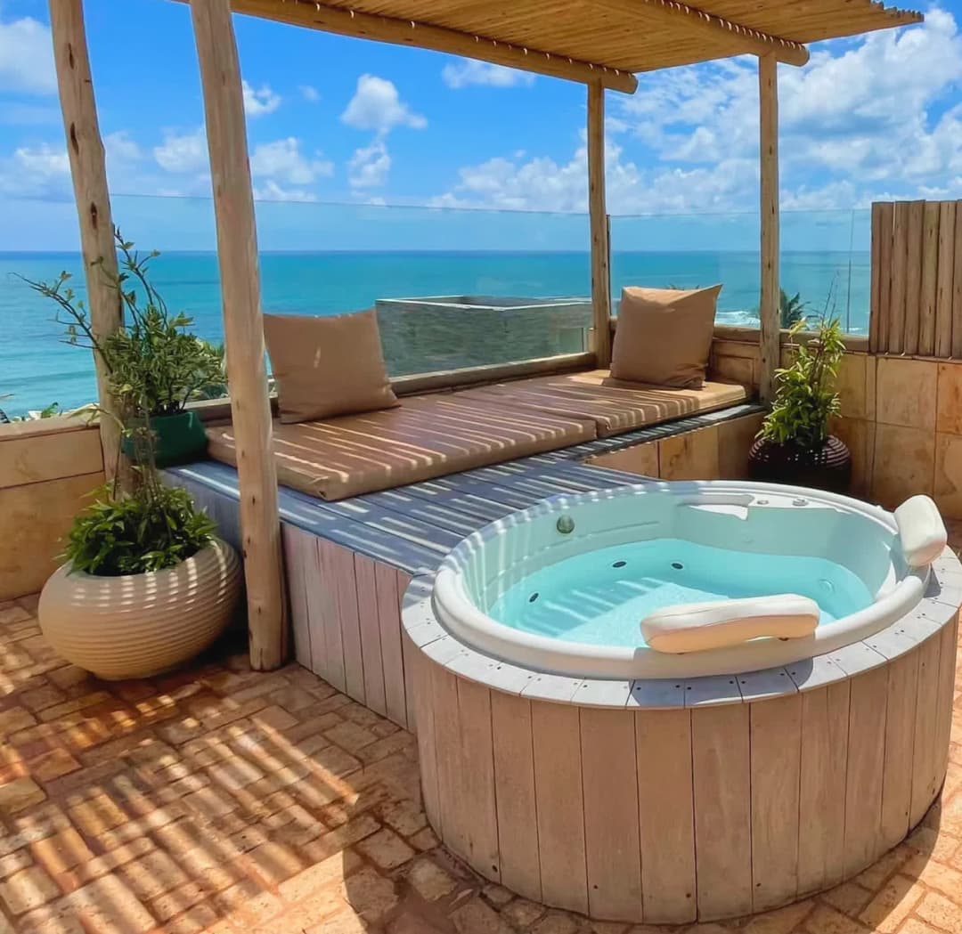 Kilombo Villas - Sibraáma Ocean - Banheira de hidromassagem circular instalada em um deck de madeira na área externa privativa. A banheira possui encostos de cabeça brancos e é cercada por uma cerca de troncos de madeira e plantas ornamentais.