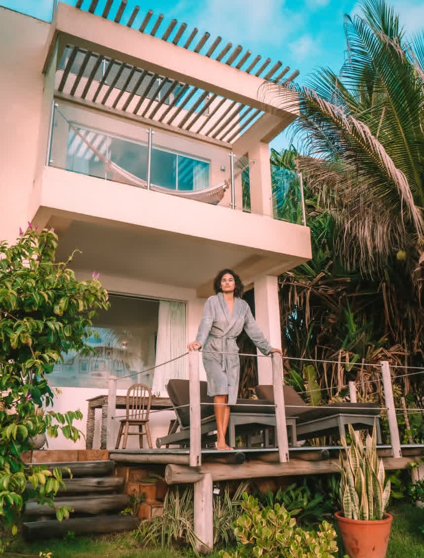 Kilombo Villas - villa Jacú Ocean - Fachada da suíte villa Jacu Ocean no hotel Kilombo Villas, destacando a arquitetura integrada à natureza com varandas de madeira e cercada por palmeiras. Uma mulher em um roupão cinza está na varanda inferior.