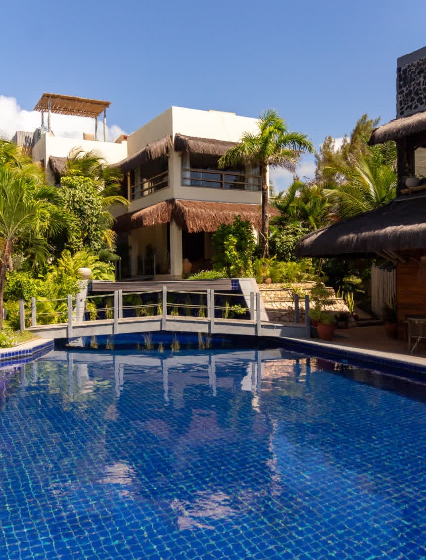 Kilombo Villas - villa Sibraáma Jardim - Vista externa da área comum do hotel, mostrando uma piscina de azulejos azuis com uma pequena ponte de madeira cinza que leva em direção à estrutura da Villa Sibraáma, cercada por palmeiras e jardins tropicais sob um céu ensolarado.