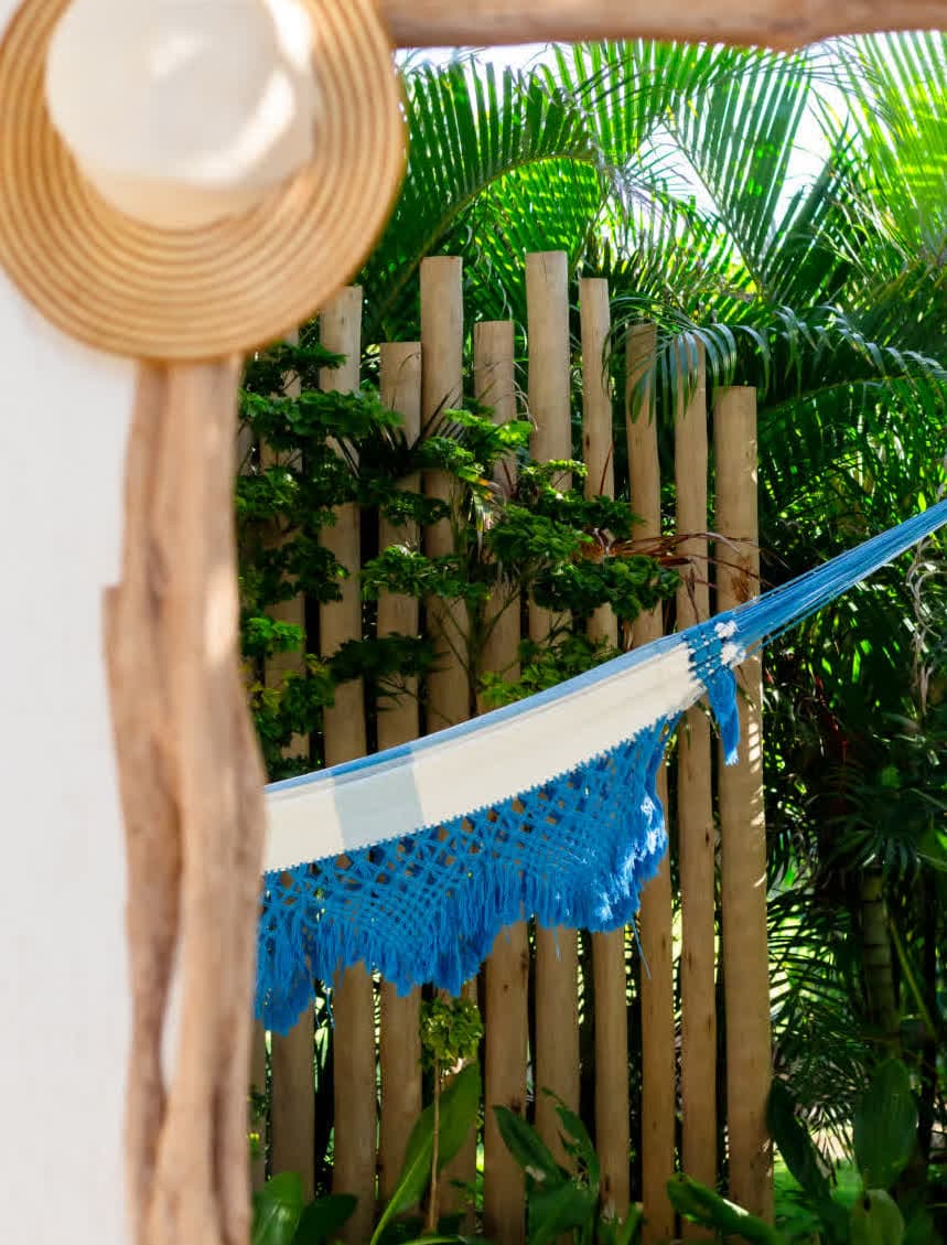 Kilombo Villas - villa Zumbi Jardim - Detalhe de uma rede de descanso em tons de azul e branco pendurada em uma área externa privativa. Ao fundo, uma cerca de troncos de madeira rústica e folhagens tropicais densas garantem a privacidade do ambiente.
