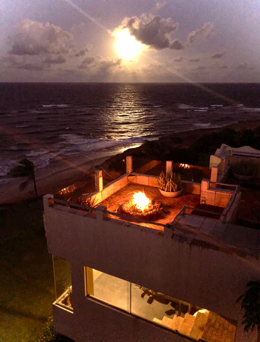 Kilombo Villas - villa Desidéria - Vista de um resort de praia ao pôr do sol com sofás na varanda, palmeiras e mar calmo, criando uma cena relaxante e paradisíaca.
