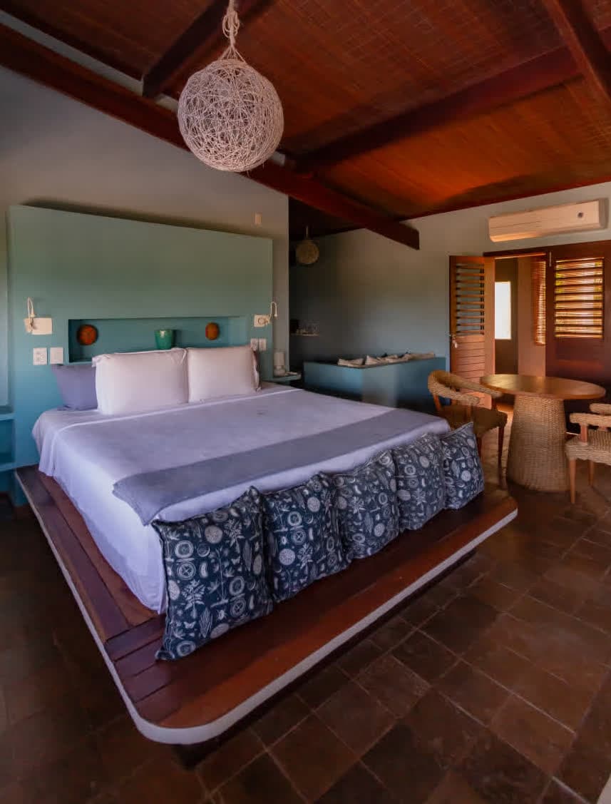 Kilombo Villas - Suíte Azul - Vista frontal do quarto da Villa Suíte Azul, destacando a cama king size sobre uma base de madeira escura e travesseiros decorativos com estampas botânicas azuis. O ambiente possui teto inclinado de madeira, piso de cerâmica rústica e uma mesa redonda com cadeiras de fibra natural.