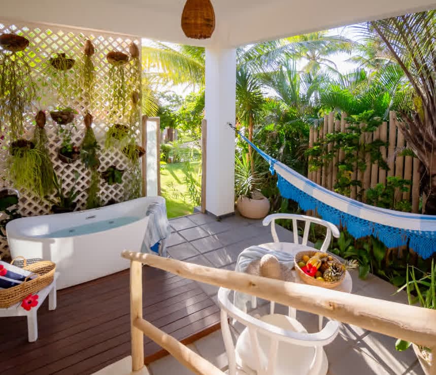 Kilombo Villas - villa Zumbi Jardim - Varanda térrea da suíte villa Zumbi Jardim com piso de cimento e um deck de madeira escura. O espaço inclui uma mesa de mármore com cadeiras brancas, uma banheira de imersão oval e uma escada que leva ao interior da villa. Uma placa antiga de sinalização de estrada decora a parede.