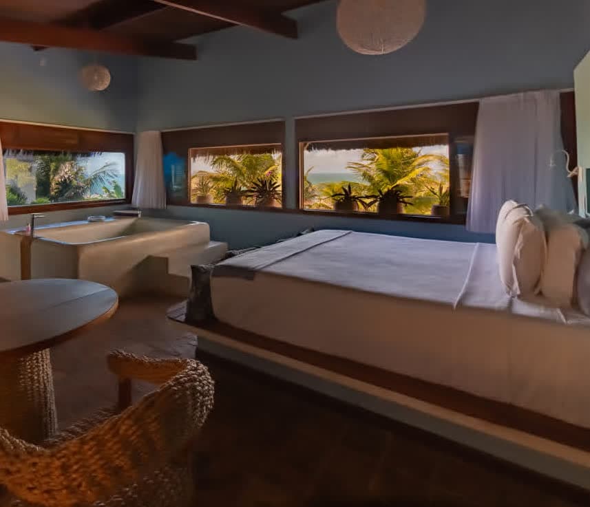 Kilombo Villas - Suíte Azul - Perspectiva da suíte integrando o quarto e a área de banho. Uma banheira de alvenaria branca com degraus de acesso está posicionada em frente a grandes janelas que oferecem vista para o jardim tropical e o mar. À esquerda, vê-se a bancada do lavatório.