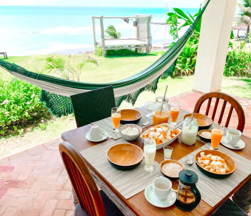 Kilombo Villas - villa Desidéria - Café da manhã ao ar livre em uma varanda com vista para a praia, incluindo sucos, ovos, frutas e café, com rede de descanso e jardim verde ao fundo.