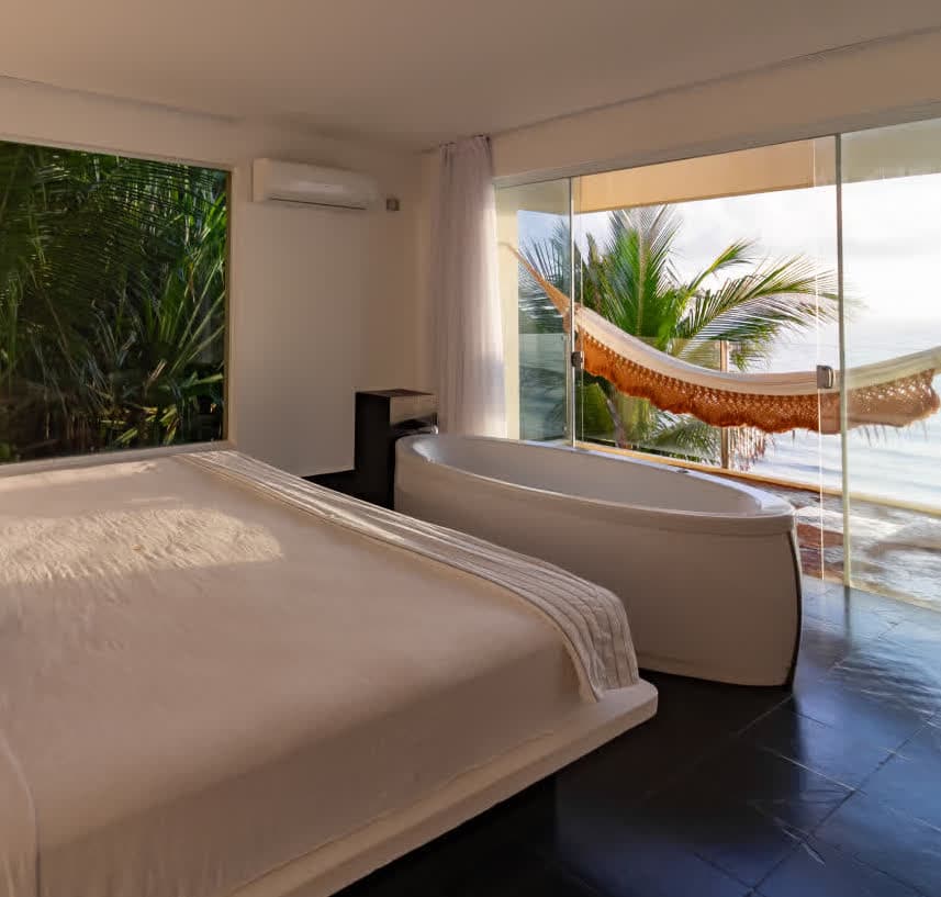 Kilombo Villas - villa Jacú Ocean - Interior da suíte master da villa Jacu Ocean, exibindo a cama king size e uma banheira de imersão estrategicamente posicionada em frente a grandes janelas de vidro com vista para o mar do Kilombo Villas.