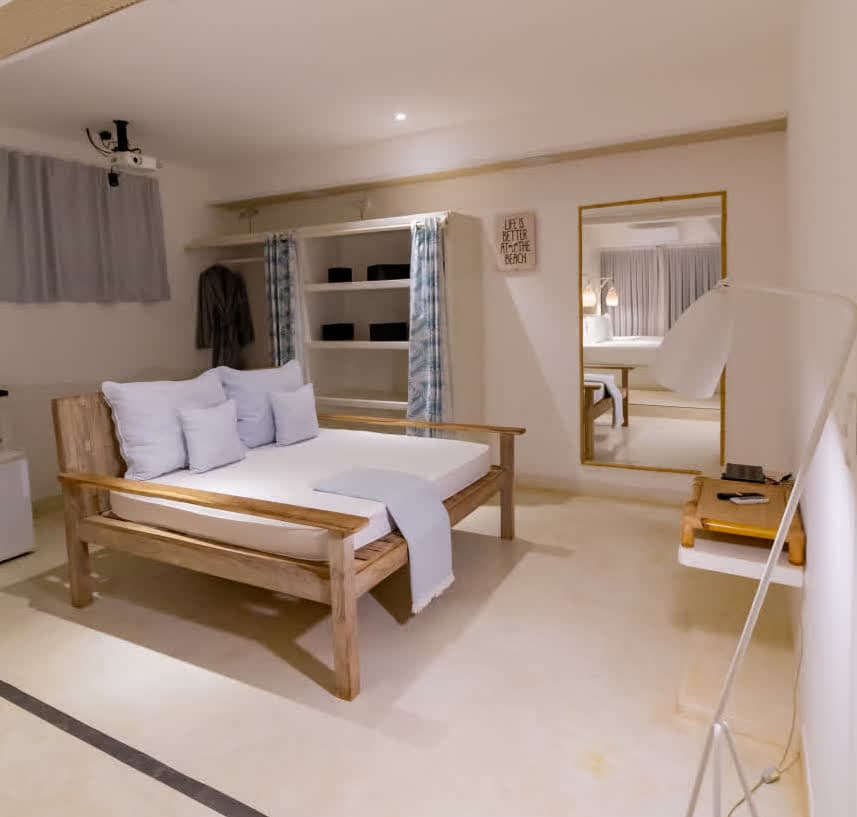 Kilombo Villas - villa Zumbi Jardim - Interior da suíte mostrando uma cama de dia feita de madeira clara com almofadas azuis. Ao fundo, um armário embutido com prateleiras brancas e caixas organizadoras pretas. Um espelho de corpo inteiro reflete a iluminação suave do ambiente.
