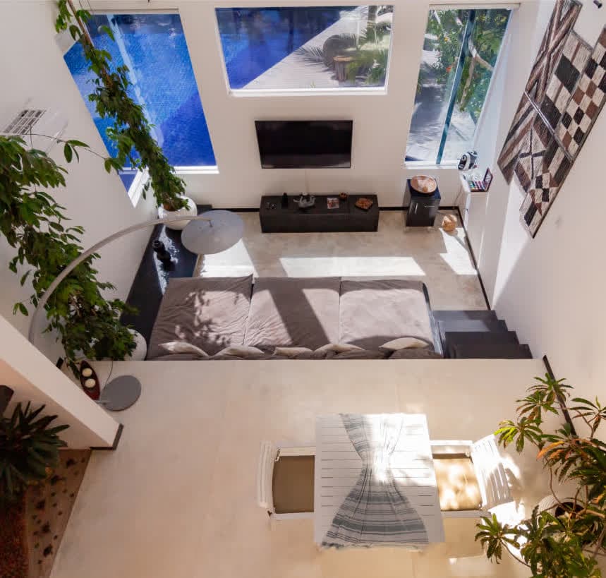 Vista superior da sala de estar de pé-direito duplo. O espaço conta com um sofá modular cinza, mesa de jantar branca e decoração que inclui arte tribal nas paredes e plantas ornamentais. Janelas altas revelam a área da piscina externa.