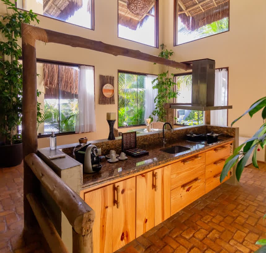 Kilombo Villas - villa Sibraáma Jardim - Espaço residencial moderno com móveis de madeira e decoração rústica. Na imagem, uma mesa de madeira clara com um candeeiro decorativo, poltrona confortável com almofada, e um abajur de fibra natural ao lado, mostrando a integração entre ambientes interno e externo.