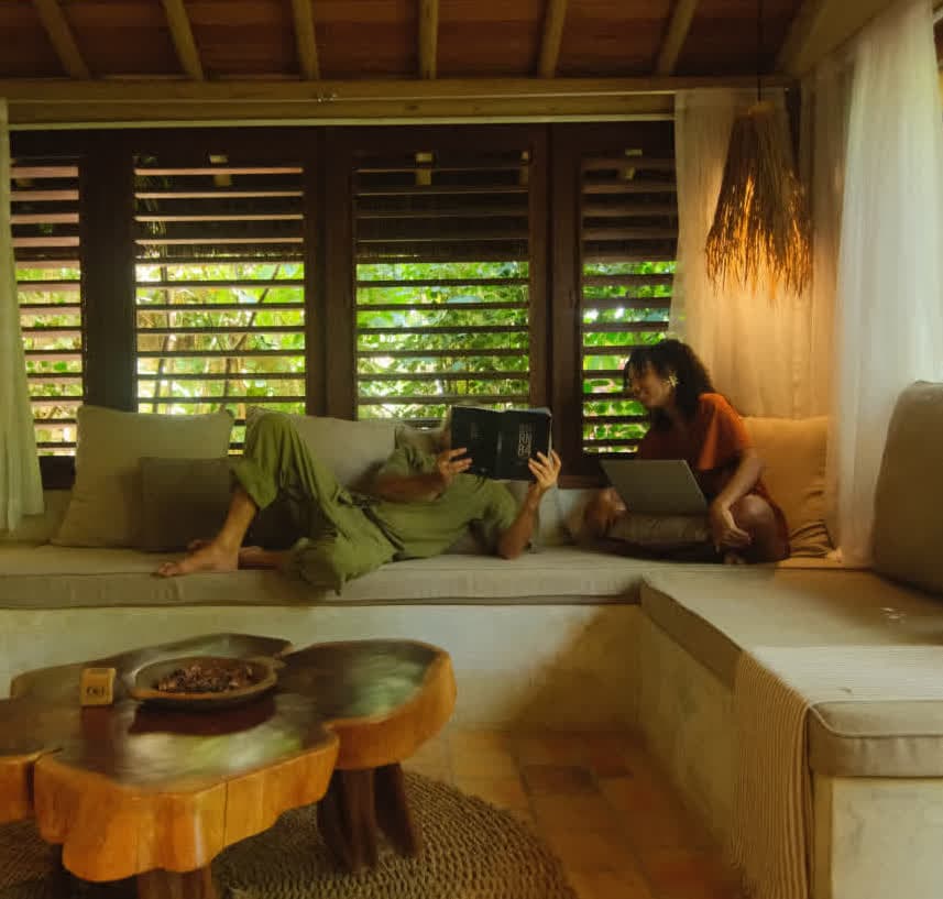 Kilombo Villas - villa Jacú Jardim - Homem e mulher usando computadores portátil e tablet, sentados em sofá confortável em sala com janelas de madeira, ambiente aconchegante e moderno.