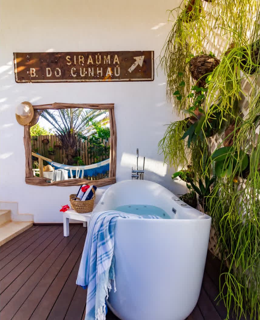 Kilombo Villas - villa Zumbi Jardim - Área externa de relaxamento com foco em uma banheira de hidromassagem branca moderna sobre o deck. A parede ao lado possui um jardim vertical em uma treliça branca com diversas plantas pendentes. Uma toalha listrada azul e branca repousa sobre a borda.
