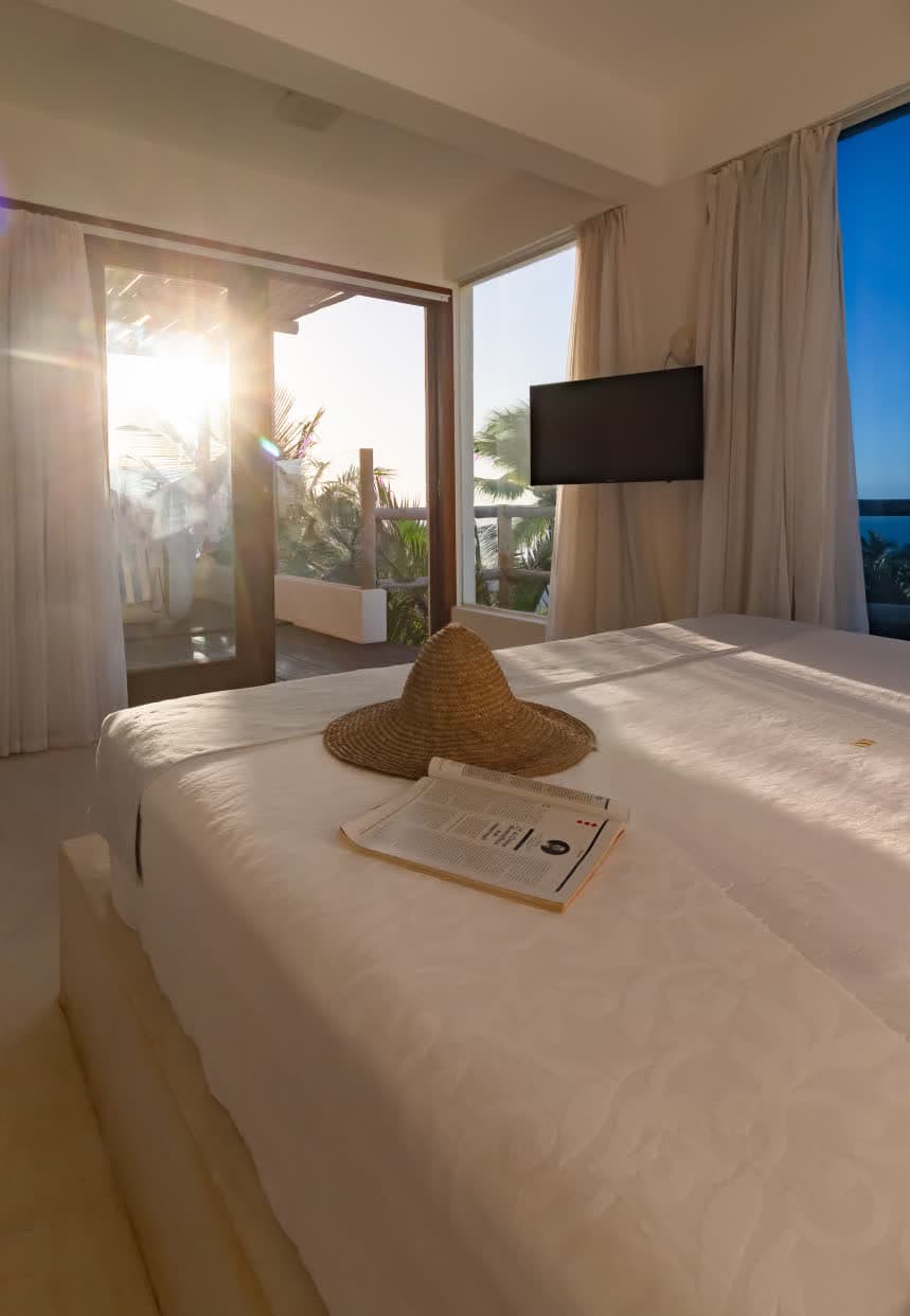 Interior do quarto durante o entardecer, com o sol brilhando intensamente através da porta da varanda. Sobre a cama branca, repousam um chapéu de palha e uma revista aberta.