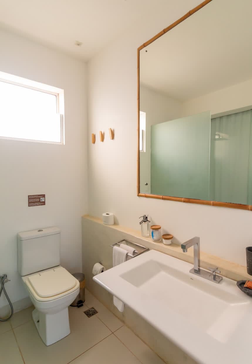 Banheiro moderno e funcional com paredes brancas. Possui uma bancada de cimento queimado com cuba de apoio branca, espelho com moldura de bambu e detalhes decorativos em madeira, mantendo a estética limpa da villa.