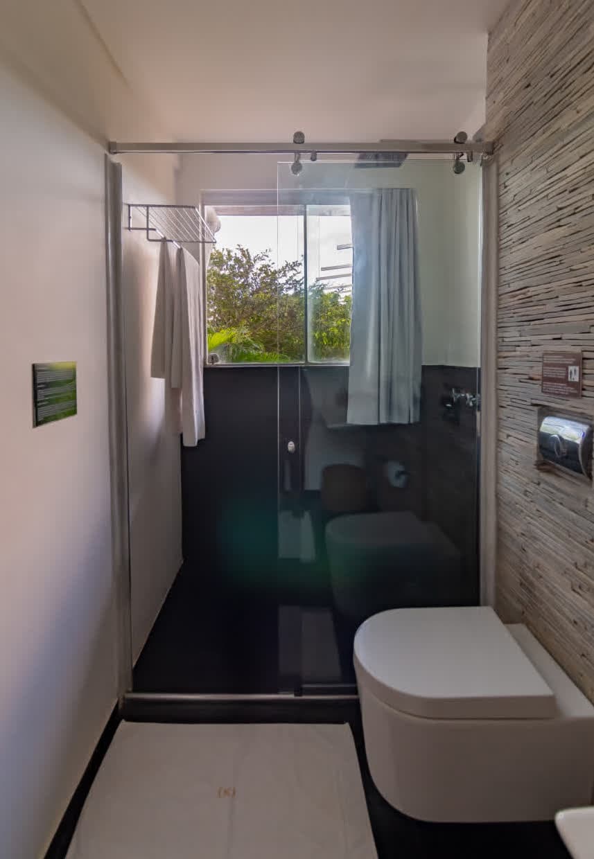 Kilombo Villas - villa Zumbi Jardim - Banheiro da suíte com design minimalista, apresentando um vaso sanitário branco suspenso e uma parede lateral com revestimento de pedras fileletadas cinzas. O box possui portas de vidro de correr e uma janela pequena que permite a entrada de luz natural.