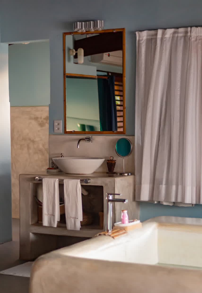 Kilombo Villas - Suíte Azul - Área do lavatório da suíte, apresentando uma cuba de apoio branca sobre uma bancada de cimento queimado. O espelho com moldura de bambu reflete o ambiente interno, e a banheira de alvenaria é visível em primeiro plano.