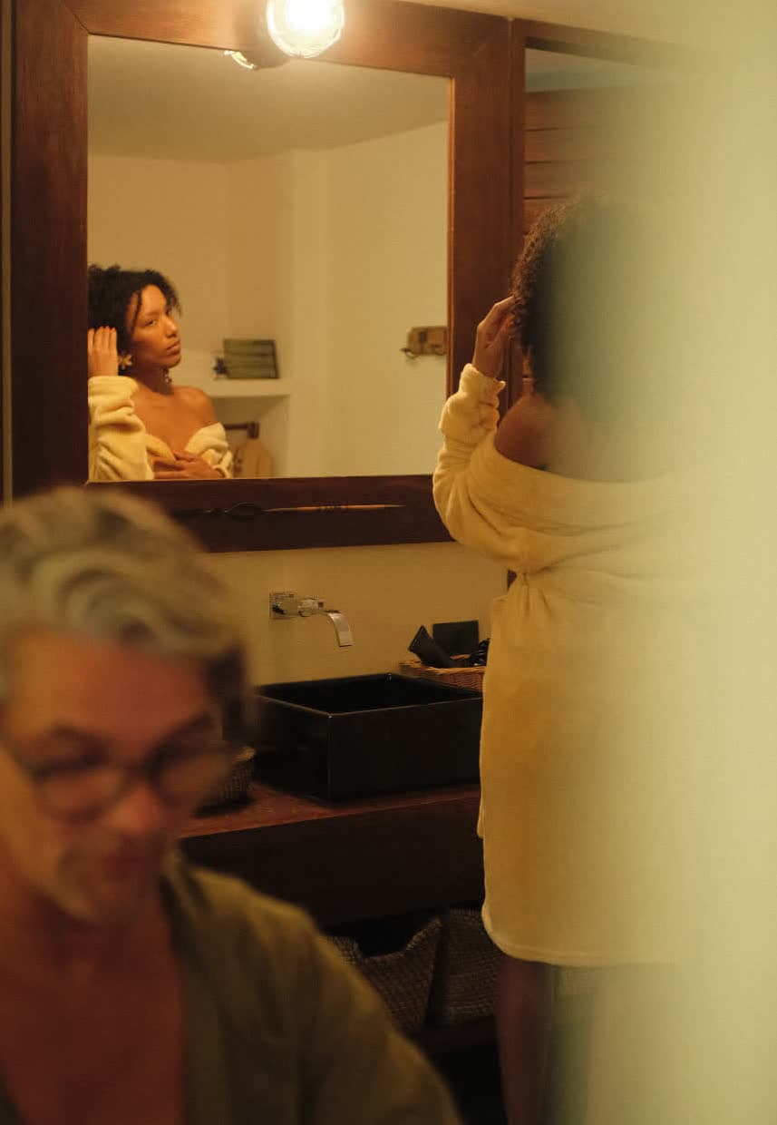 Kilombo Villas - villa Jacú Jardim - Jovem mulher de cabelo cacheado se olhando no espelho enquanto ajusta o cabelo na frente do espelho no banheiro, com um homem sorridente de óculos ao fundo.