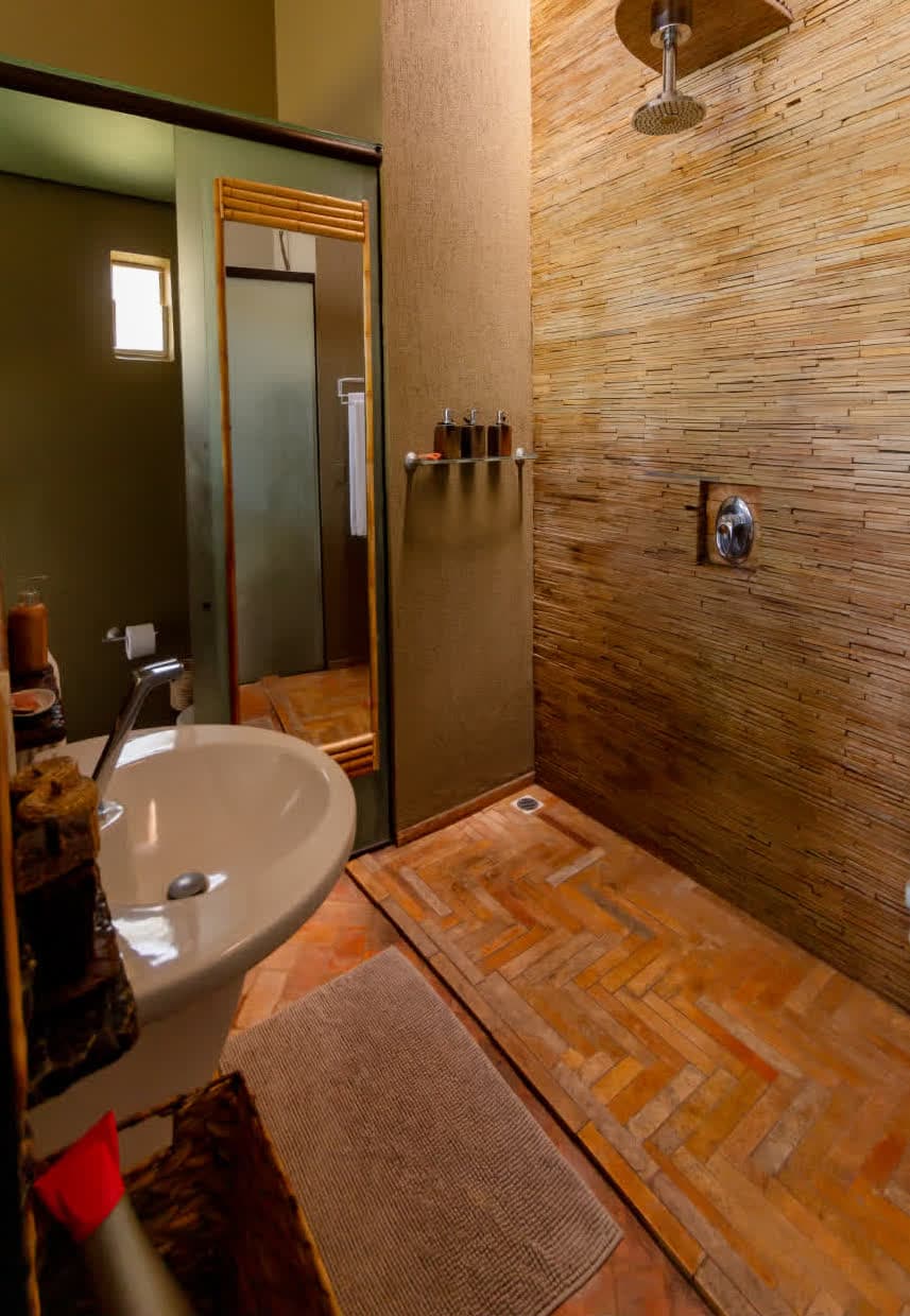 Kilombo Villas - villa Desidéria - Banheiro com parede de revestimento em madeira, ducha de teto, pia com espelho e acessórios decorativos em um ambiente aconchegante e bem planejado