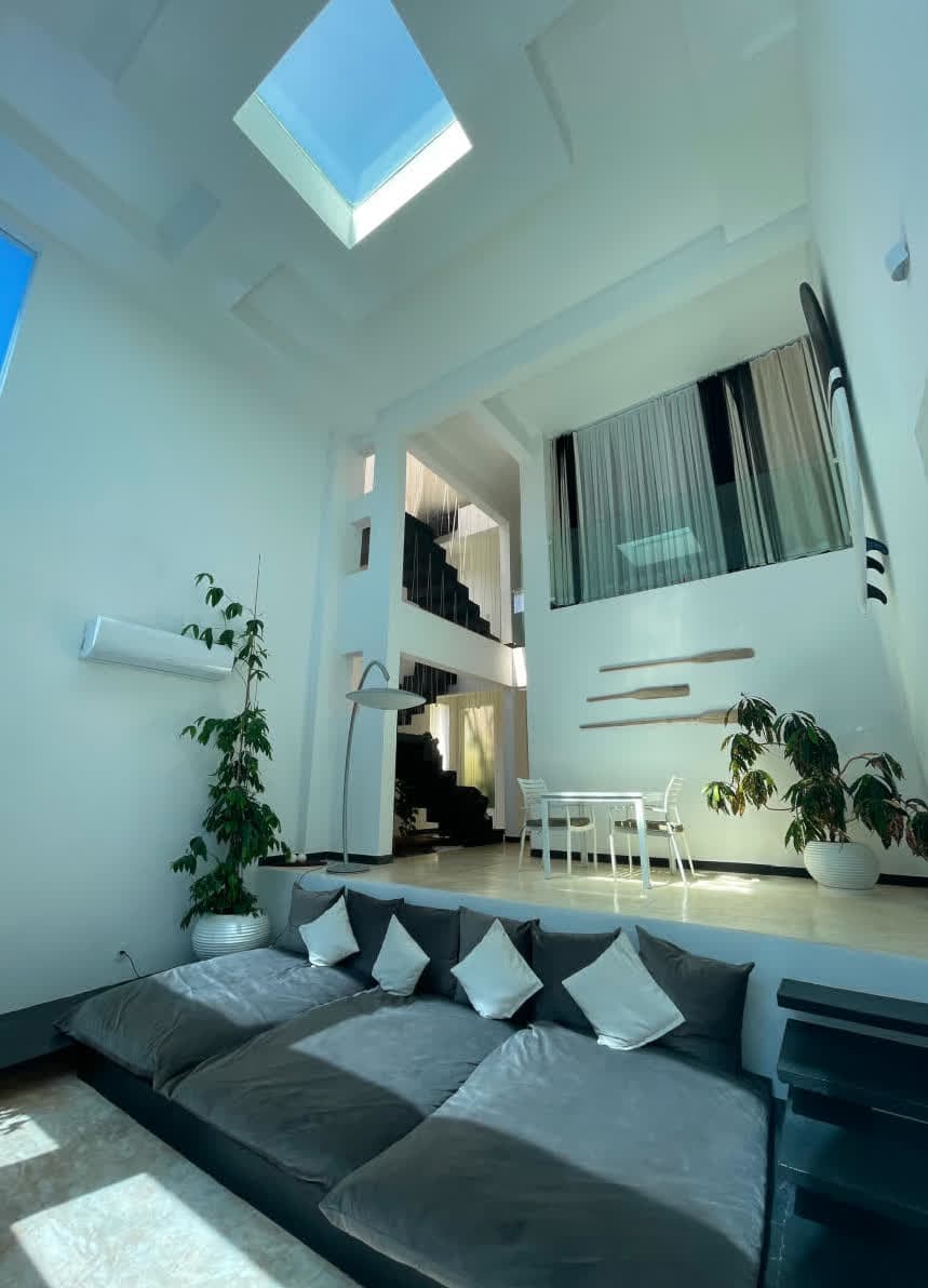 Perspectiva da sala de jantar e estar, destacando elementos decorativos únicos como uma prancha de surfe vintage e remos de madeira fixados na parede branca. A iluminação natural entra por uma abertura no teto, iluminando todo o ambiente.