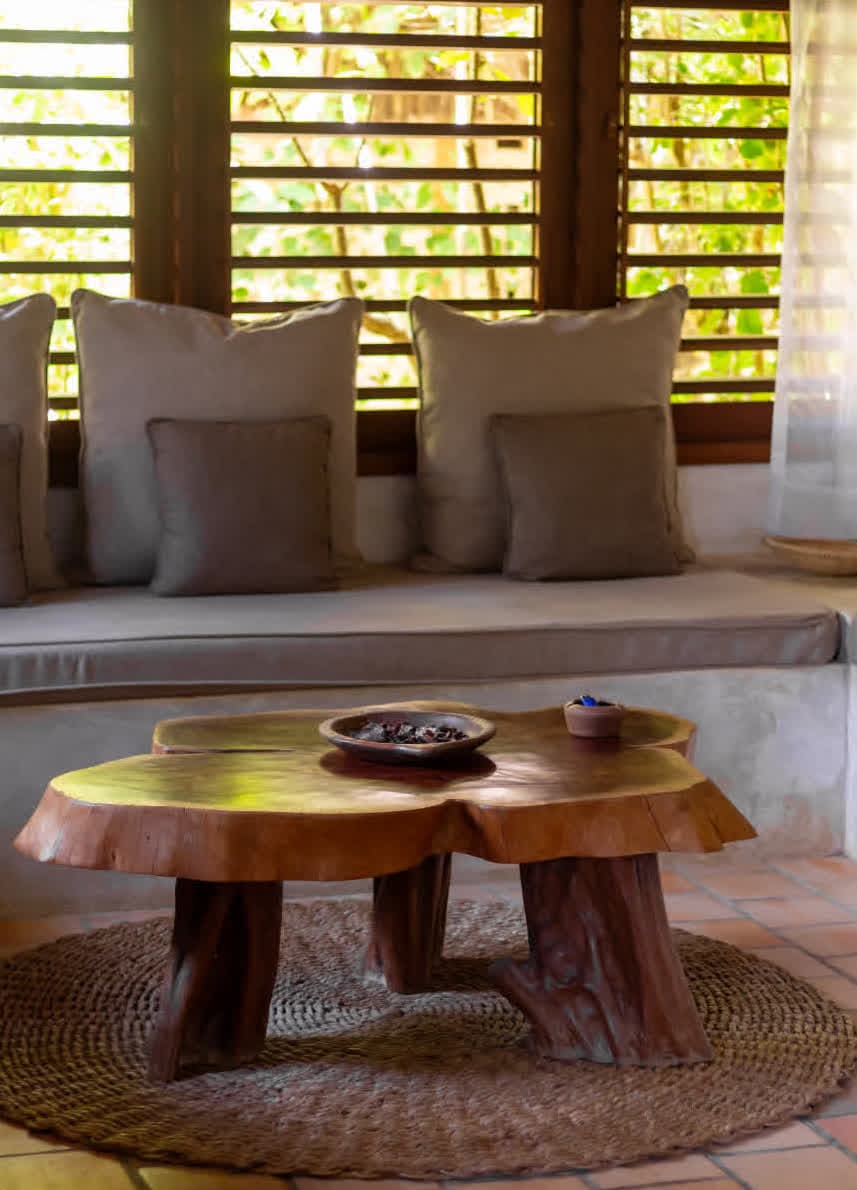 Kilombo Villas - villa Jacú Jardim - Sala de estar com almofadas bege em sofá, janela de madeira com detalhes de folhas verdes ao fundo, mesa de madeira natural, ambiente aconchegante e rústico.