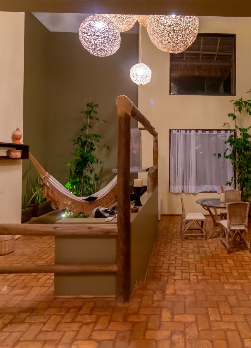 Kilombo Villas - villa Sibraáma Jardim - Quarto principal da villa Sibraáma Jardim mostrando uma cama king size decorada com uma colcha clara de textura rústica. Na parede, há uma composição decorativa de remos e um espelho com moldura de madeira e detalhes de corda. O ambiente possui piso de cerâmica rústica, uma poltrona de palha e janela com vista para o jardim.