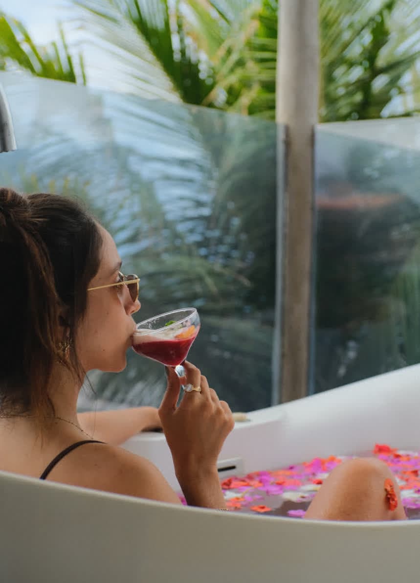 Close-up de uma mulher relaxando em uma banheira de hidromassagem repleta de pétalas de flores coloridas. Ela segura uma taça com um drink avermelhado enquanto aprecia a vista externa através do guarda-corpo de vidro.