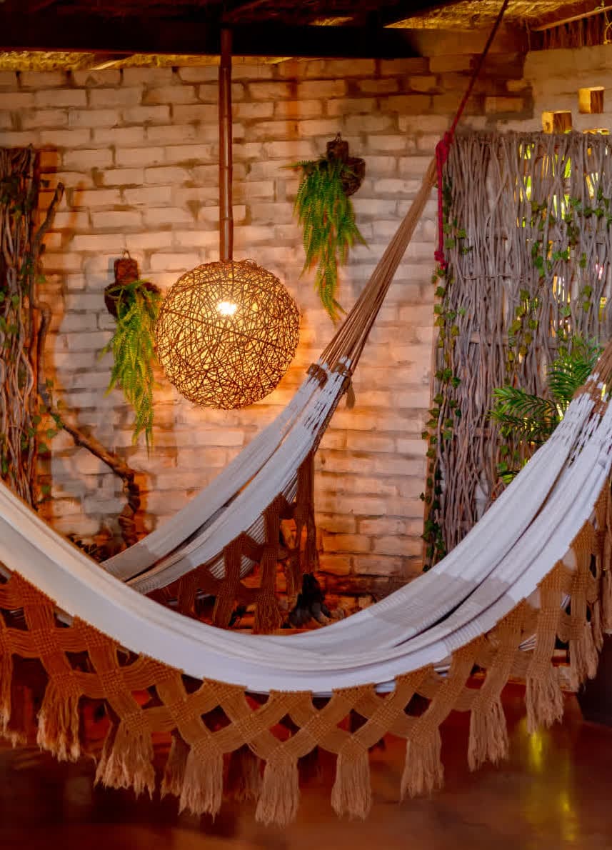 Kilombo Villas - villa Desidéria - Sala de estar decorada com rede, iluminação quente, plantas penduradas, parede de tijolos aparentes e decoração aconchegante, ideal para descanso e lazer ao ar livre.