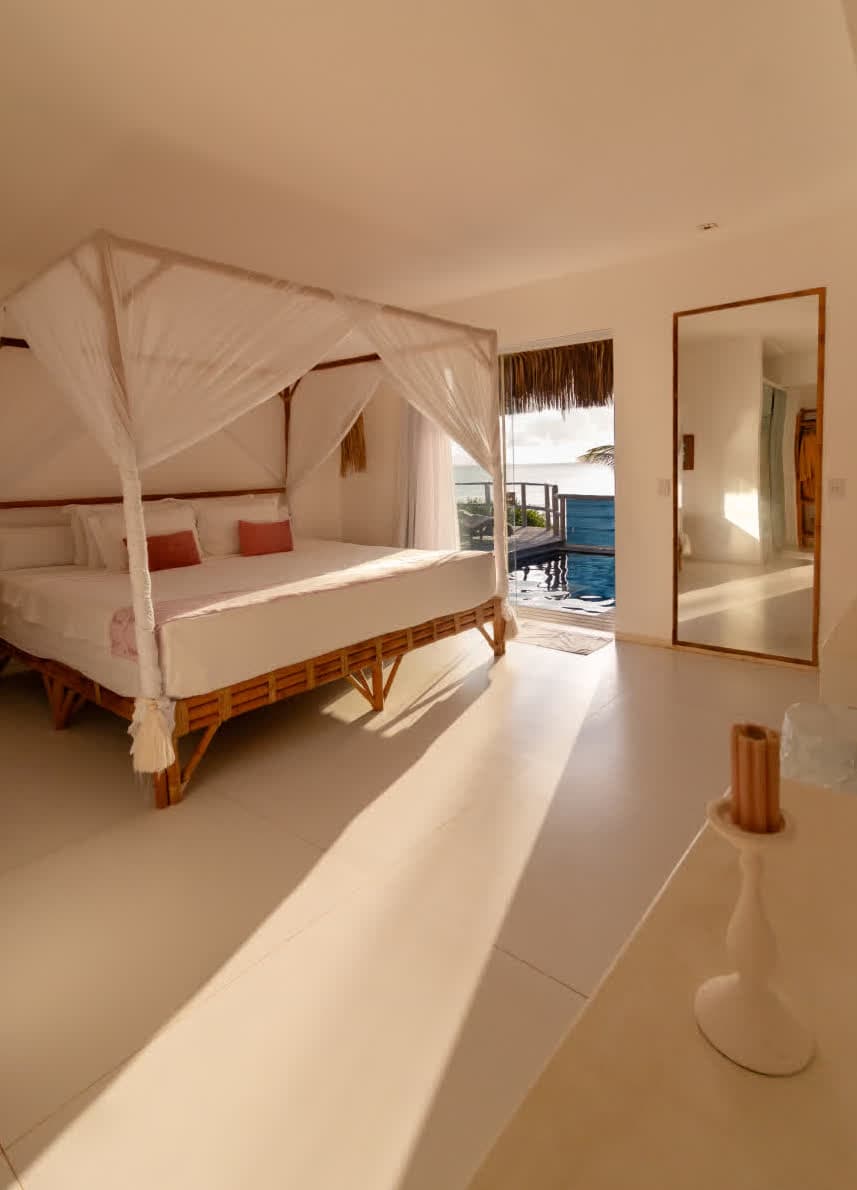 Kilombo Villas - Das Rosas - Quarto de hotel com cama de dossel, vista para a piscina e o mar ao fundo, ambiente arejado e decorado em tons neutros e naturais.