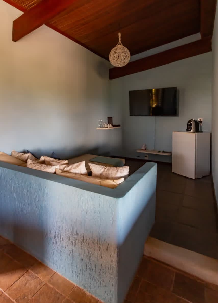 Kilombo Villas - Suíte Azul - Área de estar compacta e rebaixada da Villa Suíte Azul. O espaço conta com um sofá azul planejado, almofadas decorativas, frigobar, cafeteira e uma TV de tela plana fixada na parede azul-clara.