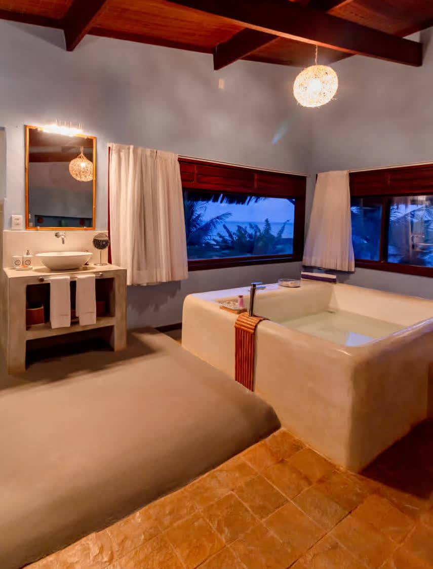Kilombo Villas - Suíte Azul - Vista noturna da área de banho e lavatório. A iluminação quente realça a textura das paredes e a água da banheira de alvenaria, enquanto as janelas ao fundo mostram o azul profundo do crepúsculo sobre o mar.