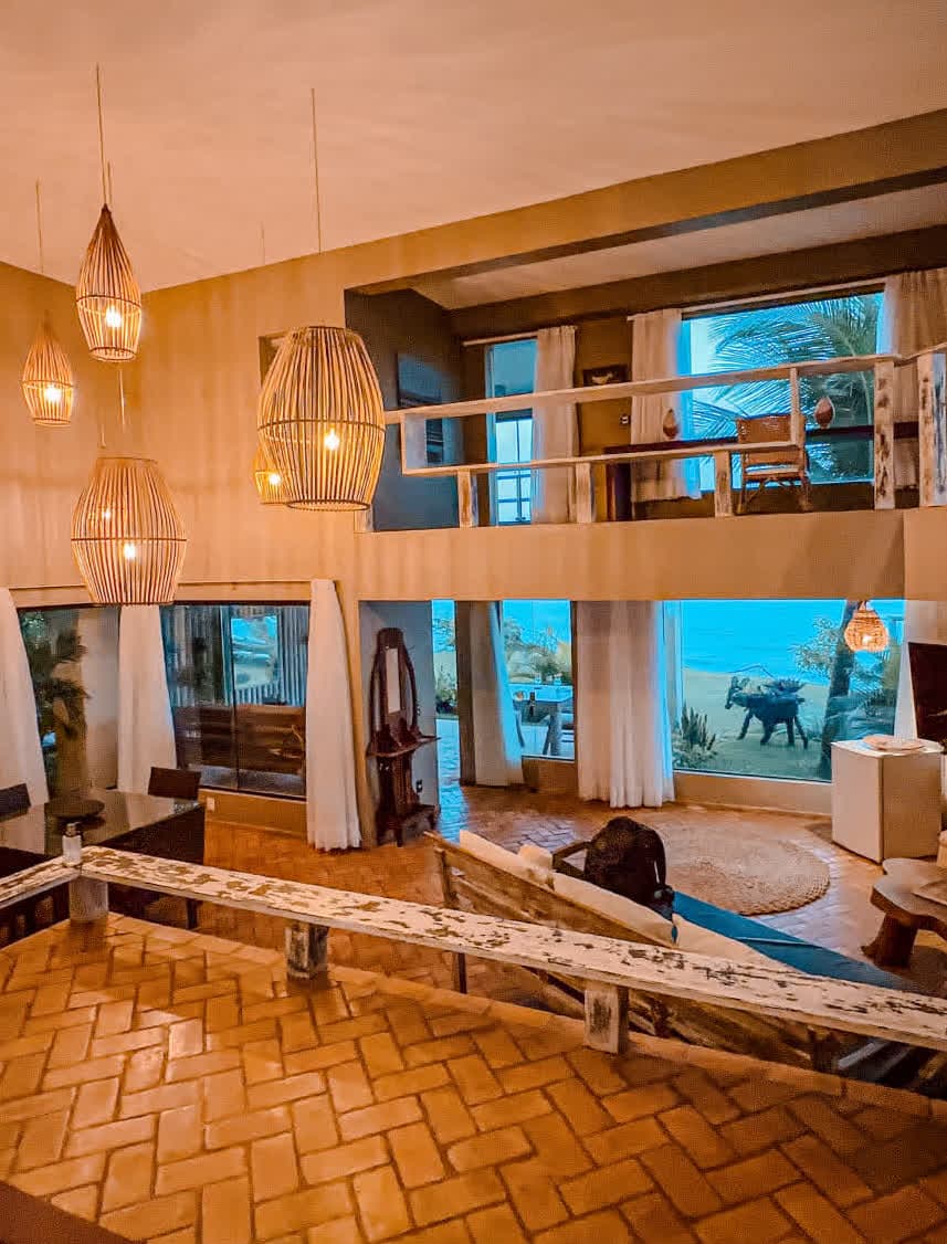 Kilombo Villas - villa Desidéria - Interior de uma sala com decoração moderna, iluminação aconchegante, janelas grandes com cortinas brancas, vista para o ambiente externo com vegetação e decoração temática.