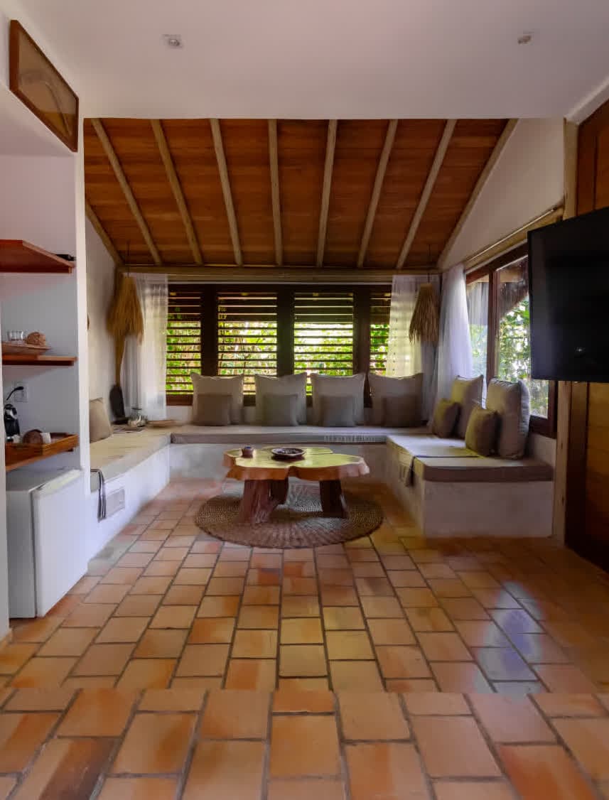 Kilombo Villas - villa Jacú Jardim - Sala de estar aconchegante com móveis de madeira, almofadas, janela com persianas horizontais, decoração rústica e ambiente bem iluminado