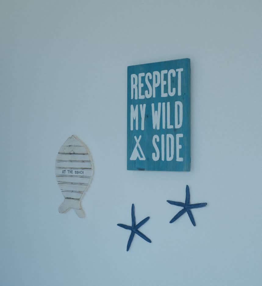 Detalhes decorativos em uma parede branca com temática marinha, incluindo uma placa azul com o texto 'Respect my wild side', um peixe de madeira com a inscrição 'At the beach' e duas estrelas-do-mar decorativas em azul escuro.