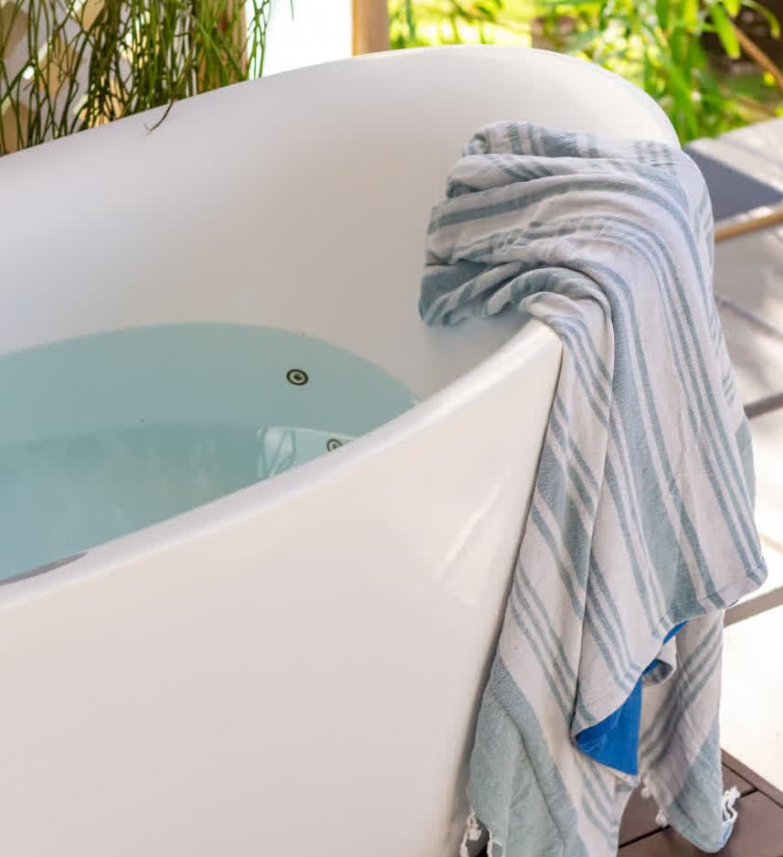 Kilombo Villas - villa Zumbi Jardim - Close-up da borda de uma banheira oval branca preenchida com água cristalina. Uma toalha de praia em tons de azul claro e branco com franjas está dobrada sobre a lateral, com o jardim ensolarado visível ao fundo.