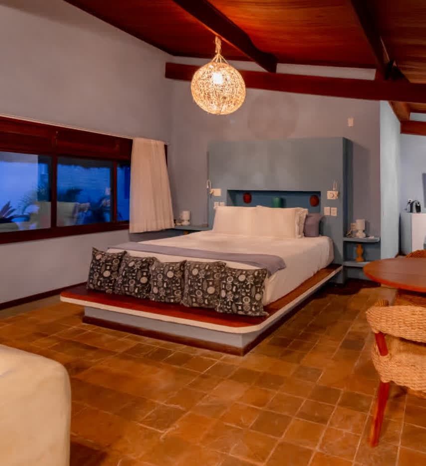 Kilombo Villas - Suíte Azul - Vista geral noturna do quarto na Villa Suíte Azul. A cama centralizada é o foco do ambiente, iluminada por um grande pendente de fibra natural que cria padrões de luz no teto de madeira e realça o tom azul suave das paredes.