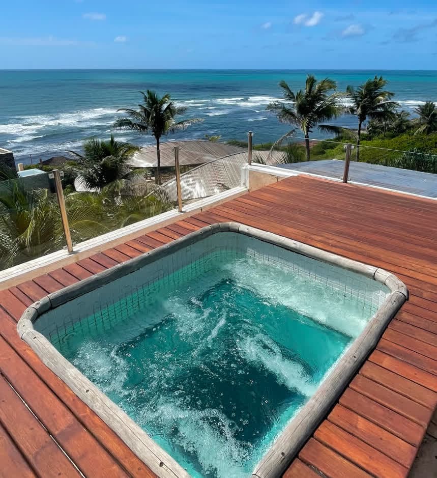 Piscina de hidromassagem quadrada em funcionamento no deck de madeira da villa Zumbi Master. A água borbulhante reflete tons de turquesa, emoldurada por uma proteção de vidro que permite a visão total da praia e das ondas do mar.