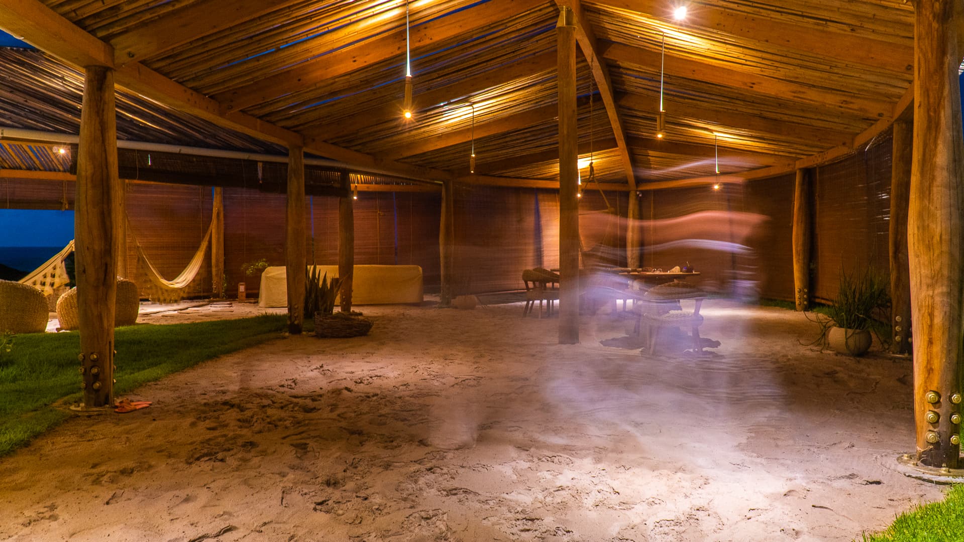 Interior rústico e iluminado do Kibo Restaurante no Kilombo Villas com areia, redes e mesas ao entardecer