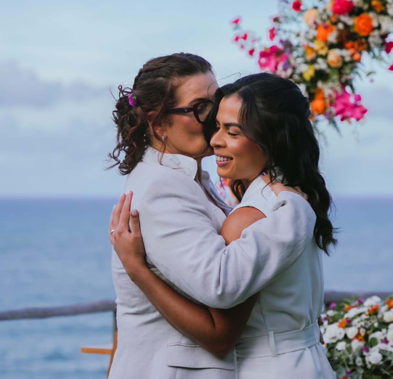 Casal se abraçando emocionado na cerimônia com arco de flores coloridas e vista para o oceano no Kilombo Villas