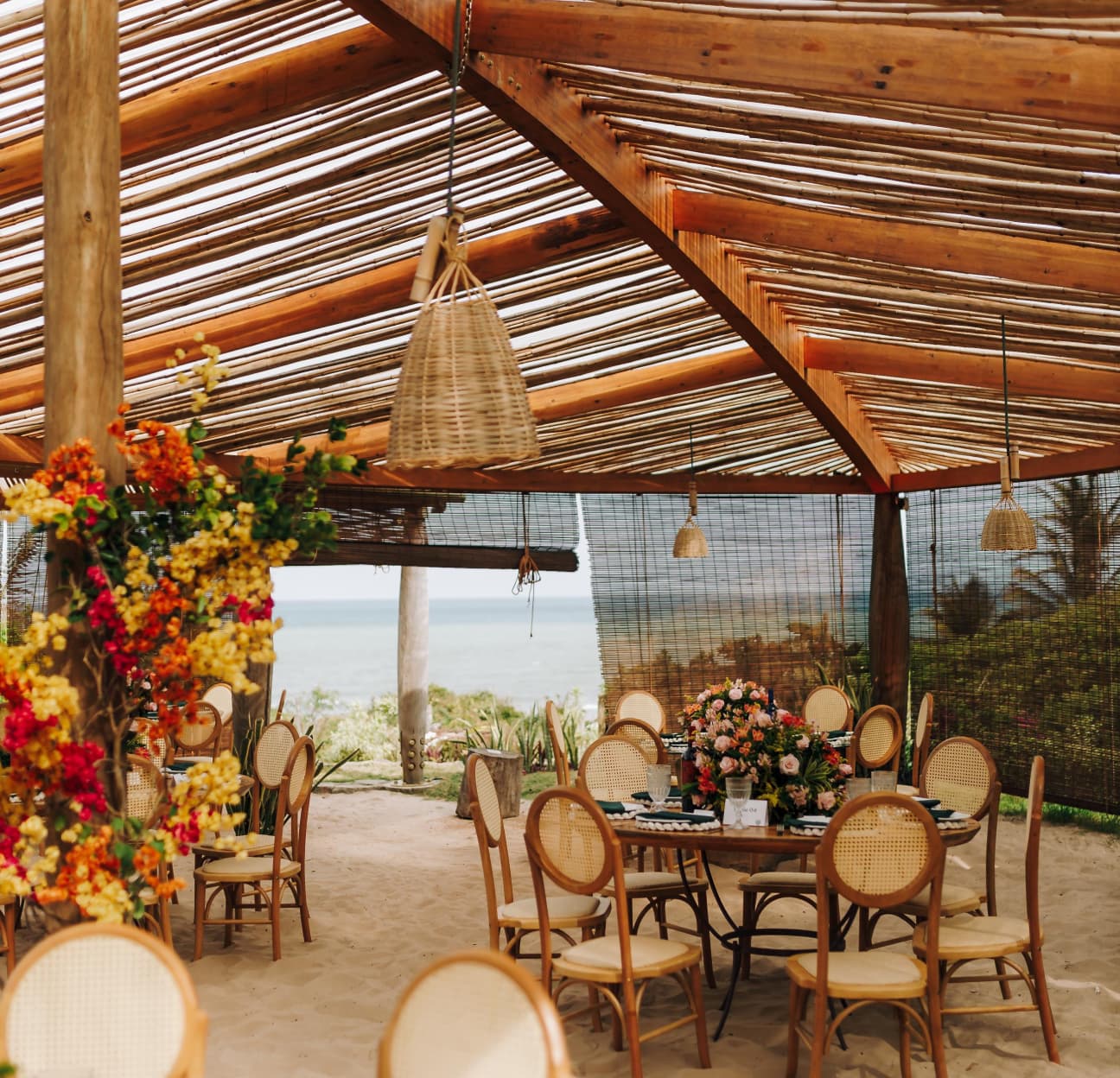 Espaço de recepção sob pergolado rústico de madeira com mesas decoradas com flores e vista para o mar no Kilombo Villas