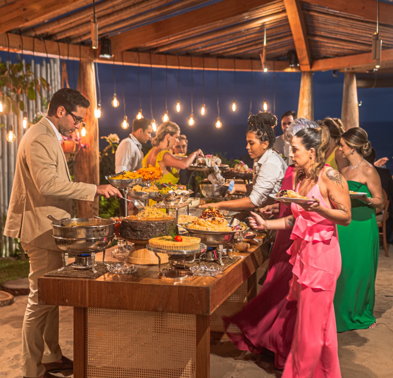 Convidados no buffet de casamento sob cordas de luzes em espaço externo ao entardecer no Kilombo Villas
